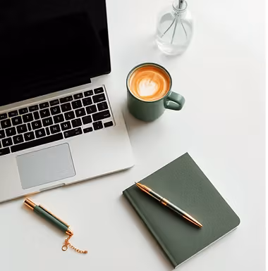 An organized desk: open laptop, green notebook, pen and a cup of coffee in cool daylight