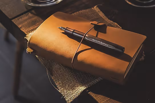 A tan leather journal tied shut with a fountain pen resting on it, warm low light
